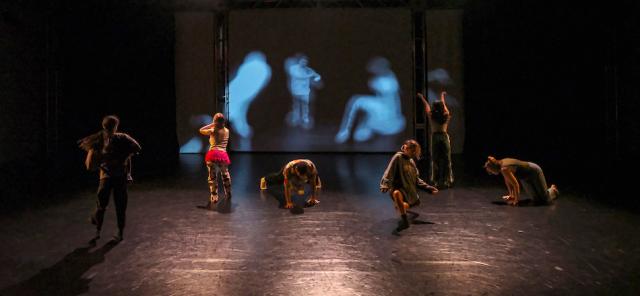 6 dancers standing, sitting, kneeling on a dimly lit stage in front of projected images of themselves that are distorted and out of focus