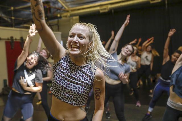 instructor with blonde hair and polka dot top reaches their right arm in the air while smiling leads a class of people in the same stretch