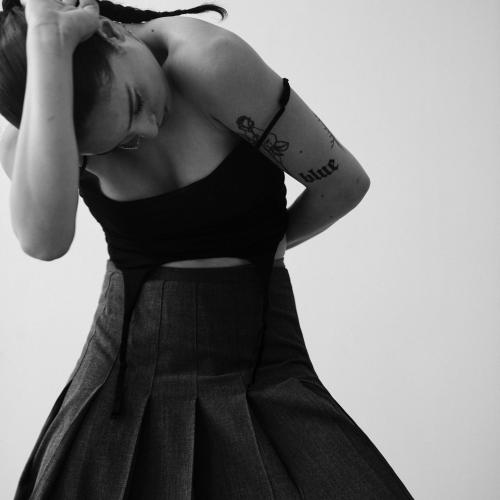 a black and white image of gigi wearing a strapped tanktop and long skirt, arched forward with their hand pressing their head down