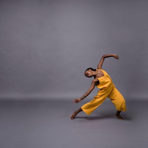 dancer in yellow jumpsuit leaning back and smiling against a grey backdrop