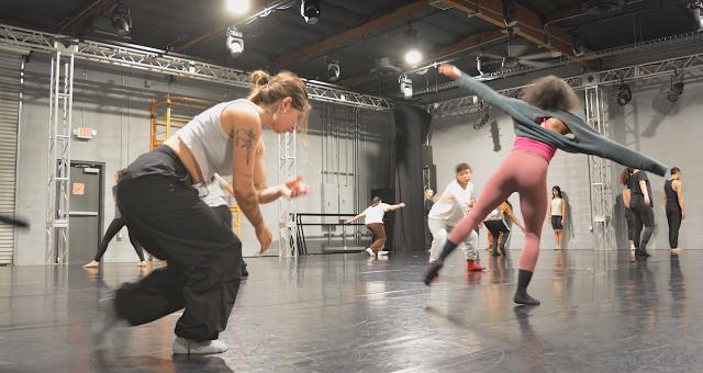 dancer crouching in black pants and grey top with brown hair next to a dancer standing on one foot reaching out to their sides with salmon colored leggings, pink top and grey longsleeve with black curly hair