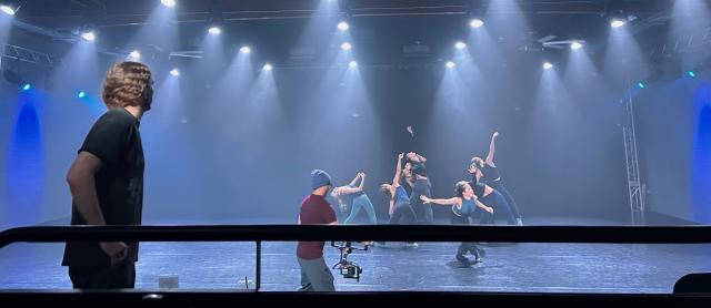 bright white lights shining down onto a black dance floor with multiple dancers in black reaching in opposing directions with a choreographer giving direction in the foreground