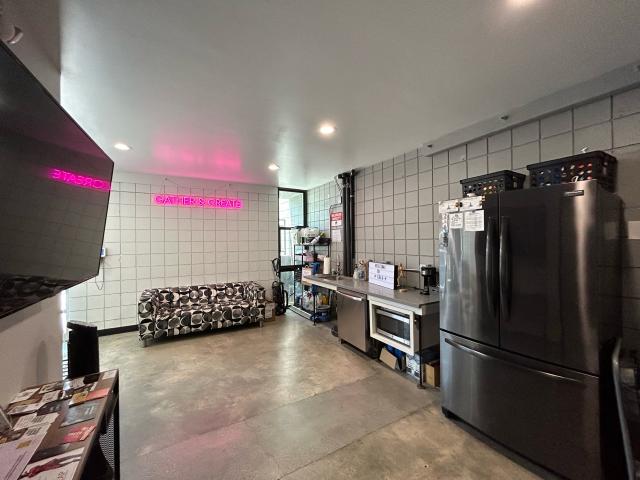 kitchen area with black refridgerator, countertop, black and white couch, pink neon reading "Gather and Create" and flat screen TV