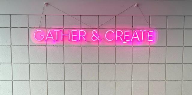 neon pink letters spelling "Gather and Create" hung on a white block wall