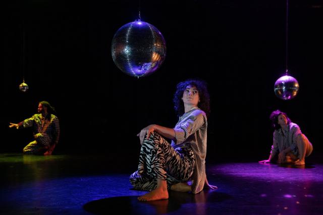 Three dancers kneel beneath disco balls in black and white costumes and purple, blue and yellow light