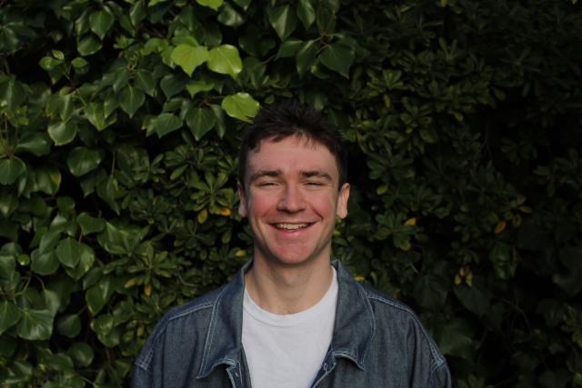 Person with brown hair wearing white shirt with a blue button down standing in front of greenery