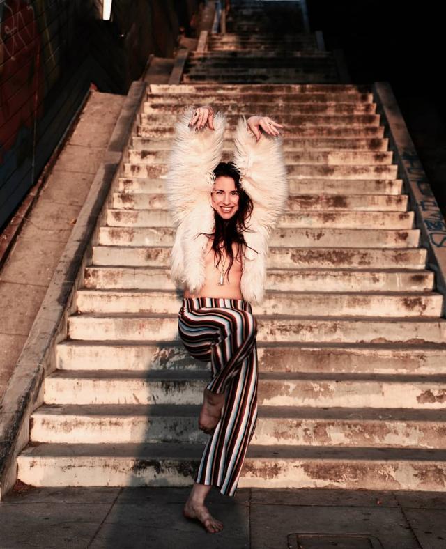 photo of a dancer wearing a fur white jacket and striped pants in front of a staircase. their leg is up to knee height and arms above their head with a joyful expression on their face