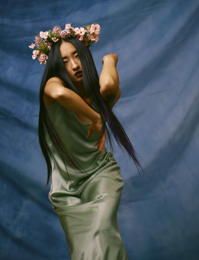 a photo of a dancer wearing a flower crown and long satin green dress with their arms folded above shoulder height. the background is a rippled blue backgorund