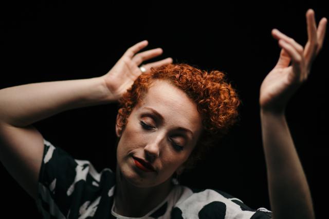 an image of a dancer with short vibrant red curly hair, with their focus down and hands placed on both sides of their head