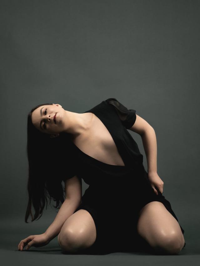 A photo of Anna Long against a dark grey backdrop. She has fair skin, and is wearing a black dress with a plunging neckline. She is placed down in a leaned over position cascading over her right ribs as her hair is draping down. Her focus is towards the camera with a bold red lip.