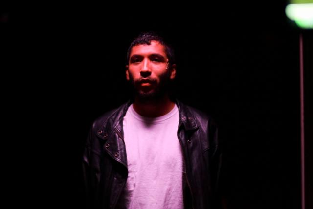 a photo of Estrellx against a dark background wearing a white shirt and faint pink lights cast uponn them