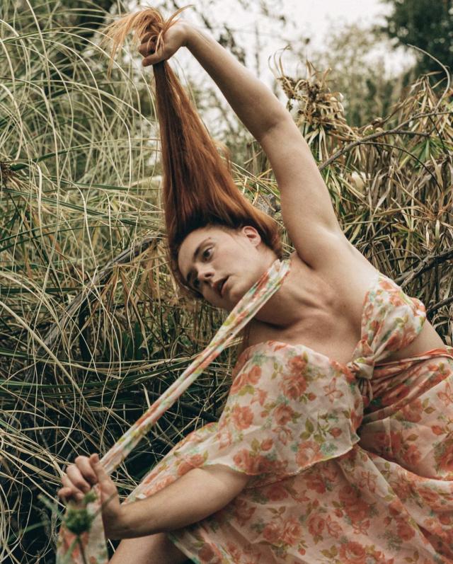 tess wearing a floral dress against green foliage, she has red hair, fair skin and is pulling both her dress string forward and hair up above her