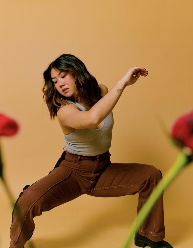 a photo of chelsea wearing brown pants and a grey top against a peachy brown backdrop. there are blurred images of roses placed in front of her.