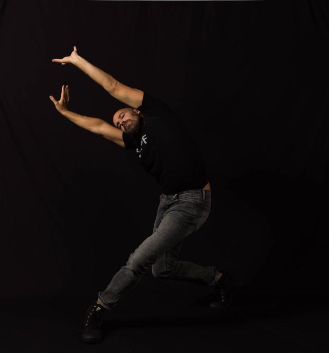 a dancer reaching both arms to the left diagonal with bent legs wearing a black tshirt and jeans on a black background