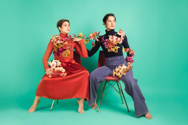 two artists seated on chairs with flowers draped on their shoulders; one wearing a red dress with short brown hair; one with blue pants and black long sleeve top with short black hair