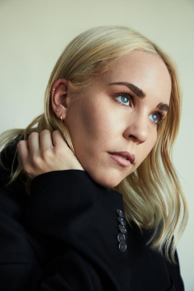 woman with blonde hair looking over her shoulder with her hand by her face wearing a black sweatshirt on a cream background