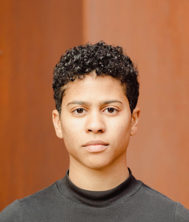 Joy stands in front of a brown background. She is wearing a dark grey top, has short curly hair and is staring directly into the camera.