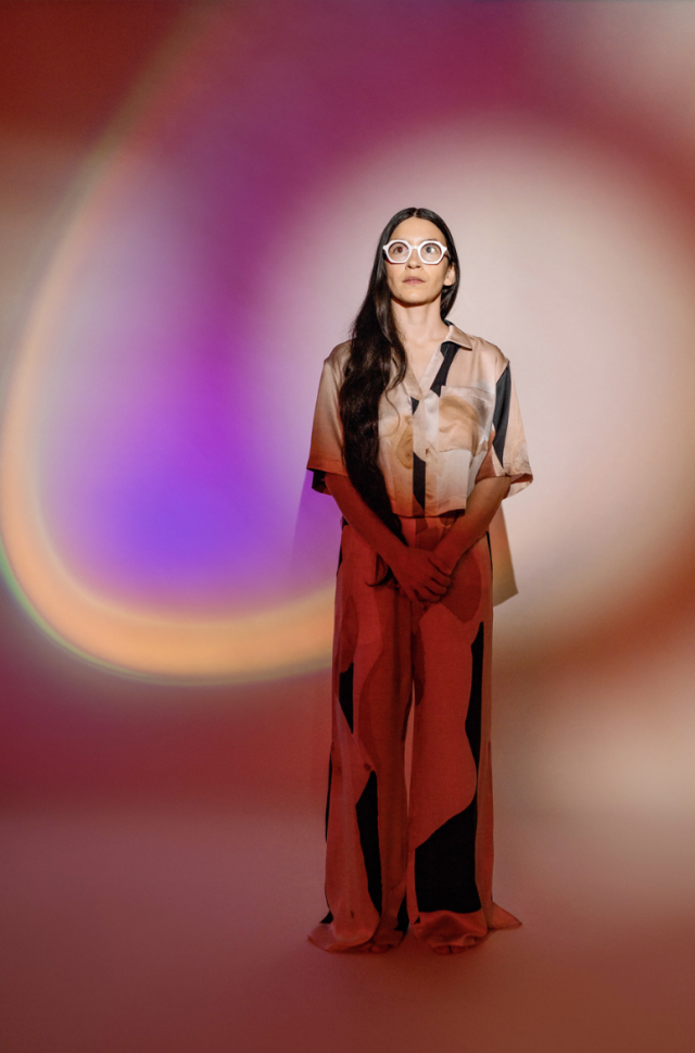 a photo of teresa standing with long dark hair and glasses looking into the distance, she is cast with different colors against her and the wall behind her