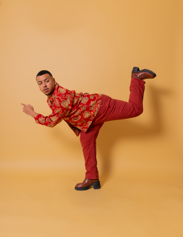 image of derrick against a peachy brown backdrop. they are wearing all red with a vintage rug fabric jacket and brown boots. their positioned in an back attitude leaning forward and looking backward