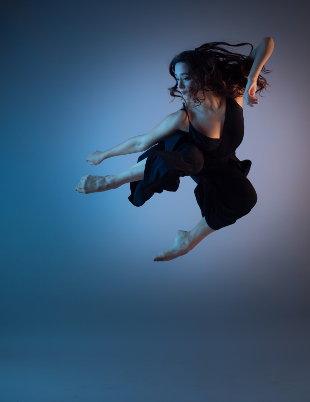 a photo of a dancer at the height of a jump with their legs narrowed towards the side, the background is a blend of muted blues and pinks