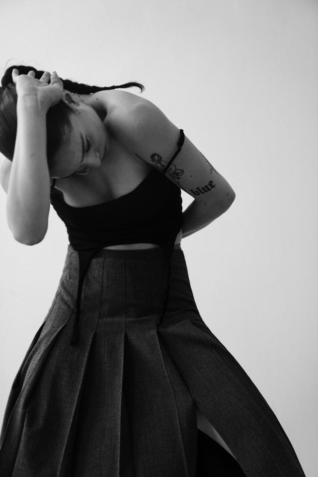 a black and white image of gigi wearing a strapped tanktop and long skirt, arched forward with their hand pressing their head down