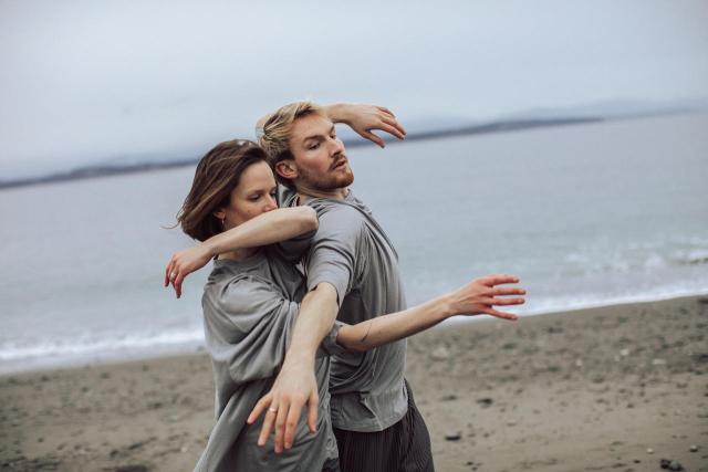 a photo of two dancers intertwined in movement on the sand of a overcast beach