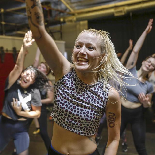 instructor with blonde hair and polka dot top reaches their right arm in the air while smiling leads a class of people in the same stretch