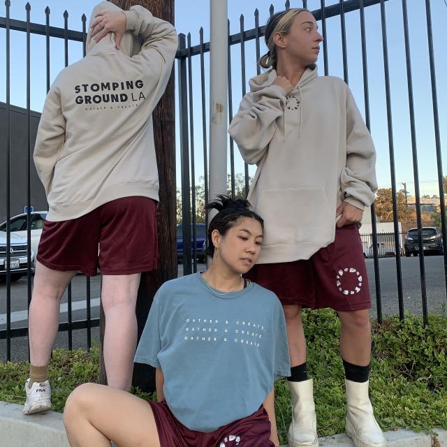 Three figures standing at a black fence with cream sweatshirt, red shorts, and blue shirt with the SGLA logo