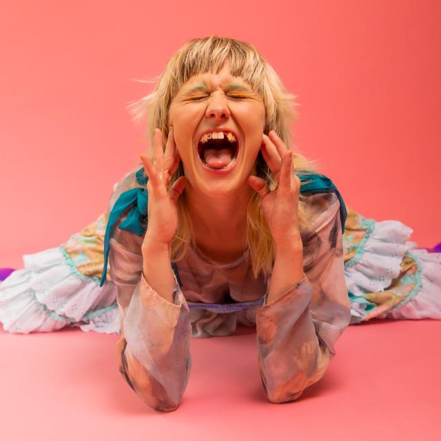 girl laying on stomach with elbows up holding face, mouth open, missing tooth pink backdrop