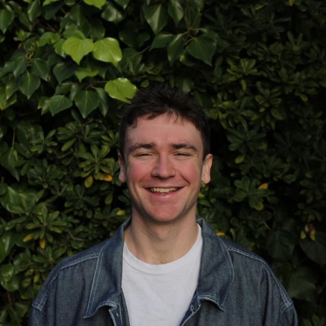 Person with brown hair wearing white shirt with a blue button down standing in front of greenery