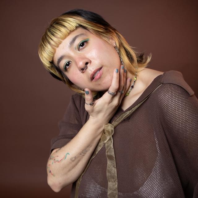 Dancer looking into camera holding their cheek wearing a brown top against a brown backdrop