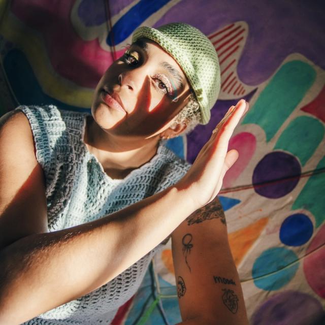 dancer holds left hand up to their face wearing a green beanie, knit top and standing in front of a colorful painted backdrop