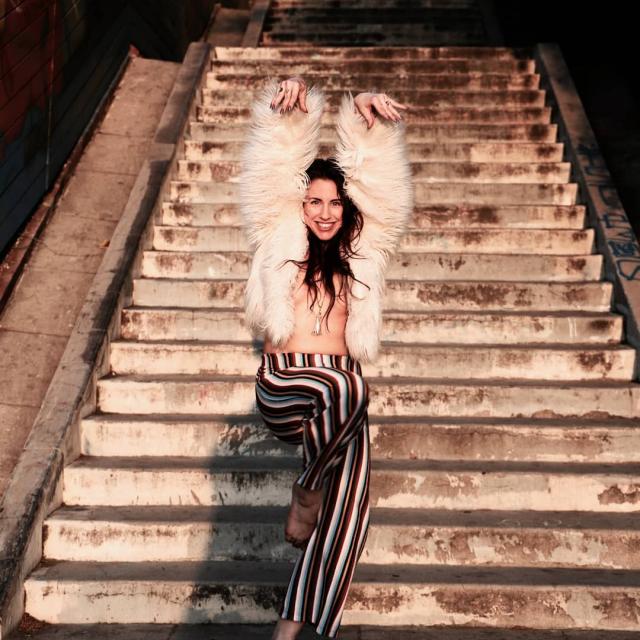 photo of a dancer wearing a fur white jacket and striped pants in front of a staircase. their leg is up to knee height and arms above their head with a joyful expression on their face