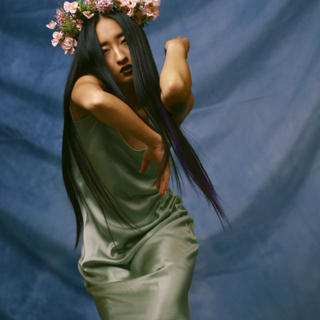 a photo of a dancer wearing a flower crown and long satin green dress with their arms folded above shoulder height. the background is a rippled blue backgorund