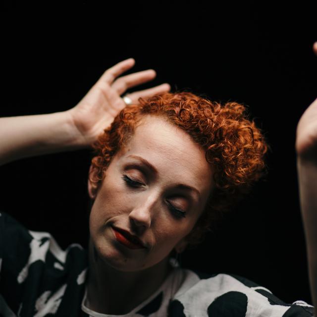 an image of a dancer with short vibrant red curly hair, with their focus down and hands placed on both sides of their head