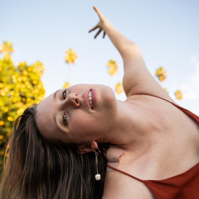 dancer with long blonde hair leaning to the side and reaching towards the sky with yellow flowers and a red dress