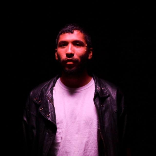a photo of Estrellx against a dark background wearing a white shirt and faint pink lights cast uponn them