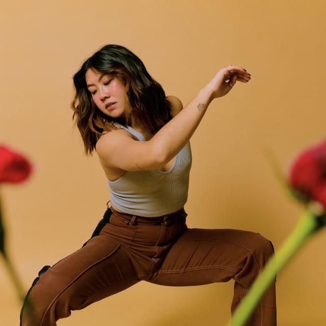 a photo of chelsea wearing brown pants and a grey top against a peachy brown backdrop. there are blurred images of roses placed in front of her.