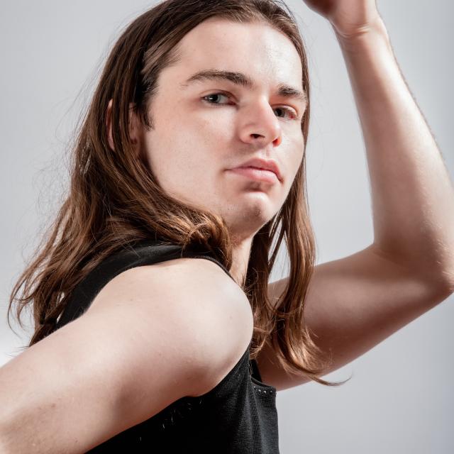 a photo of Will against a grey background with long brown hair positioned with their hand above their head and focused into the camera