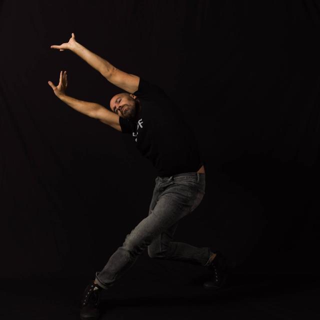 a dancer reaching both arms to the left diagonal with bent legs wearing a black tshirt and jeans on a black background