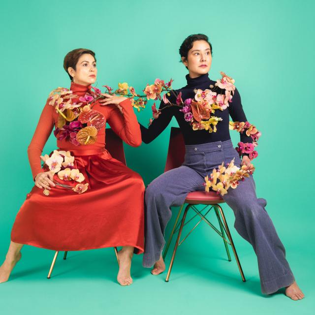 two artists seated on chairs with flowers draped on their shoulders; one wearing a red dress with short brown hair; one with blue pants and black long sleeve top with short black hair