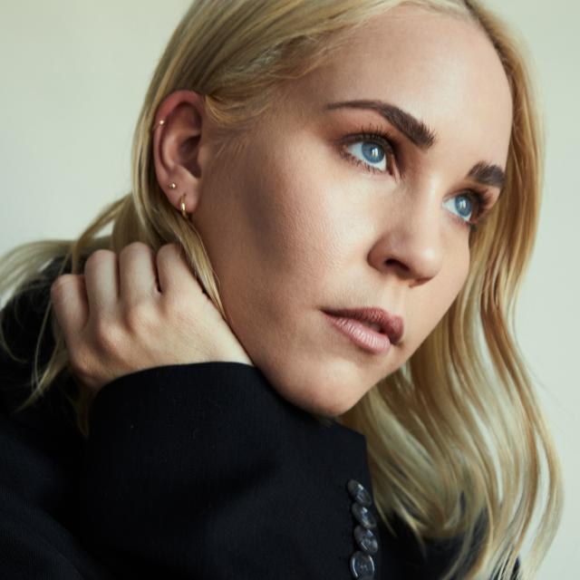 woman with blonde hair looking over her shoulder with her hand by her face wearing a black sweatshirt on a cream background