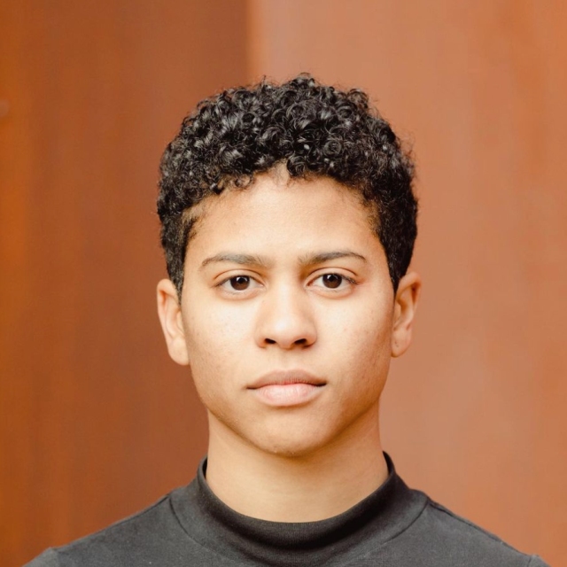 Joy stands in front of a brown background. She is wearing a dark grey top, has short curly hair and is staring directly into the camera.