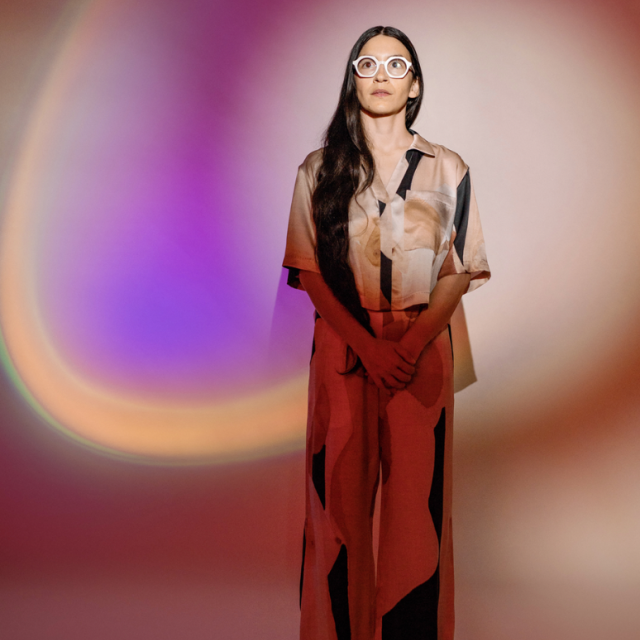 a photo of teresa standing with long dark hair and glasses looking into the distance, she is cast with different colors against her and the wall behind her