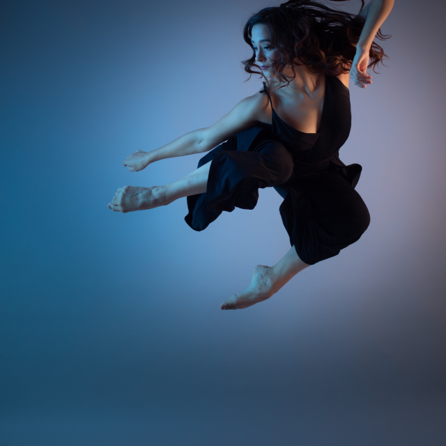 a photo of a dancer at the height of a jump with their legs narrowed towards the side, the background is a blend of muted blues and pinks