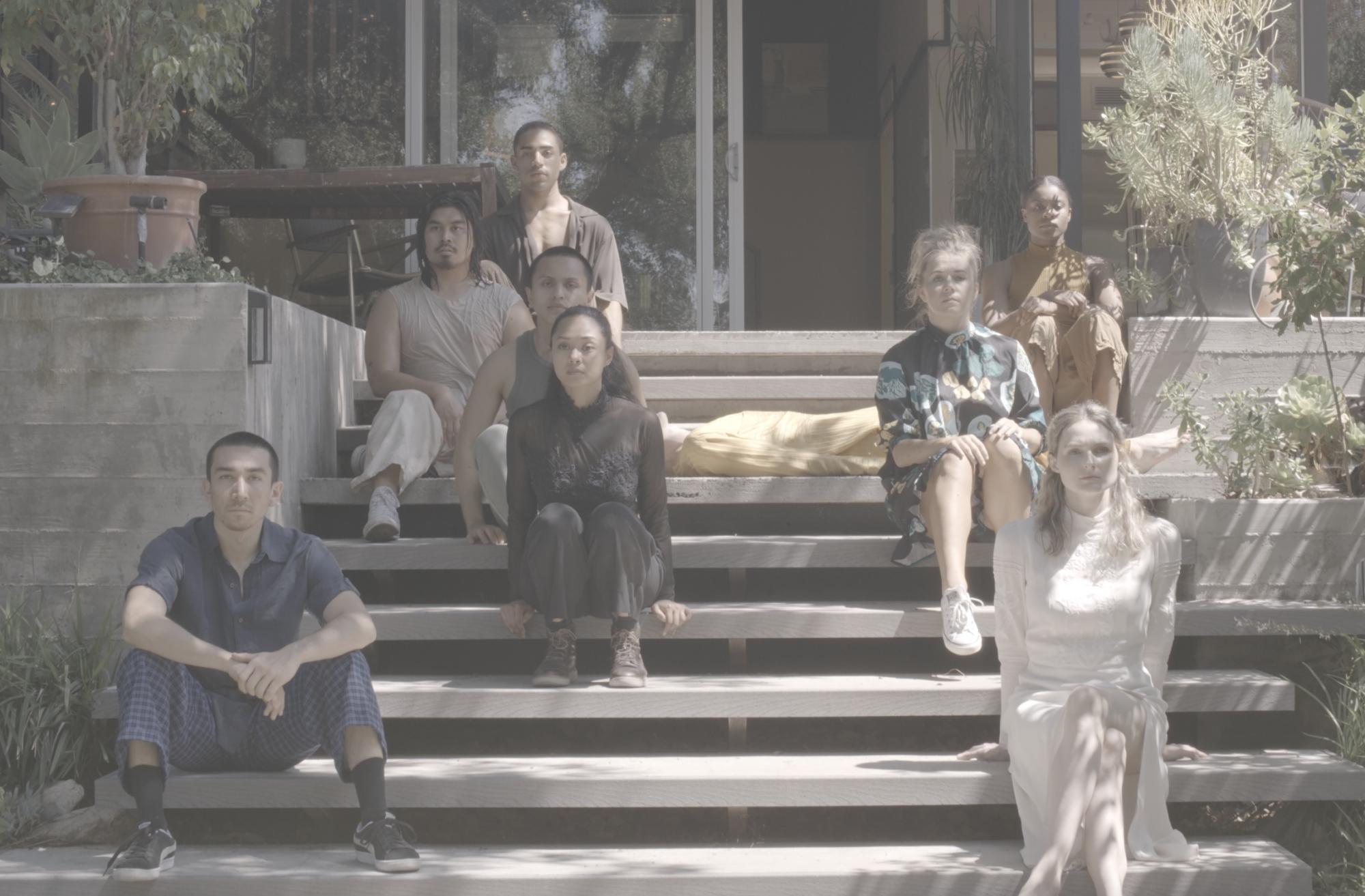 wide shot of 8 people sitting on a set of outdoor concrete stairs, with an additional person laying down, mostly obscured by the other bodies. The 8 people look out past the camera. The colors and tone of the photo feel muted and a bit nostalgic with mostly neutrals and earth tones. They are surrounded by foliage