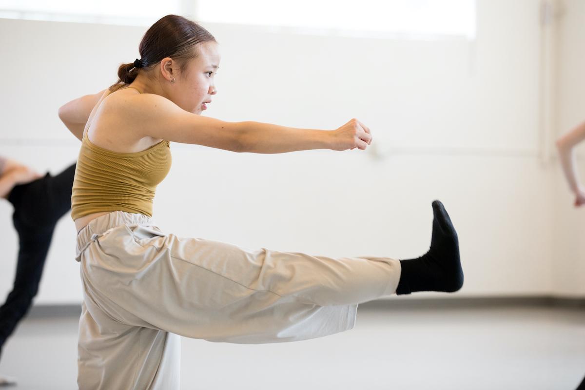 an image of a dancer wearing cream pants, black socks and a mustard top arched forward with their leg at a 90 degree angle