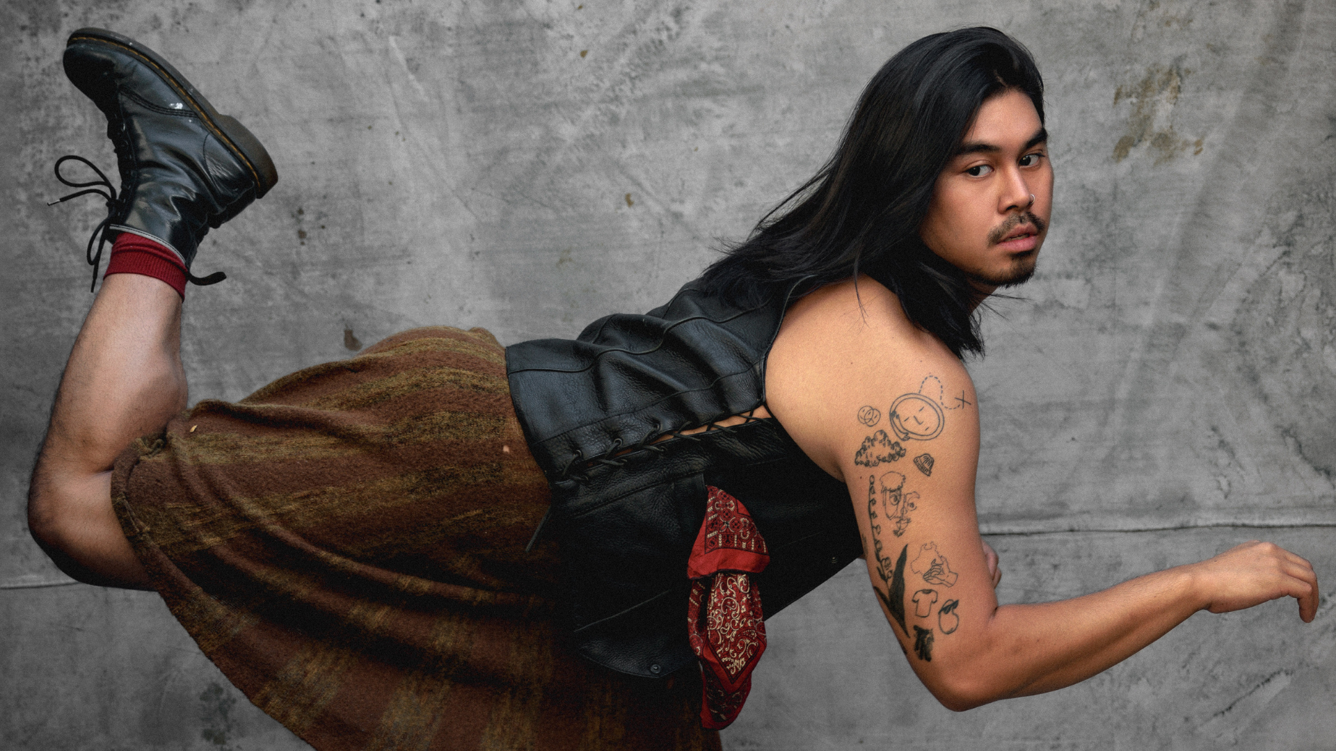 a photo of a dancer with brown skin, long black hair and facial hair is pitched over with their back leg in attitude. they are wearing a leather vest and a subtle patterned skirt.
