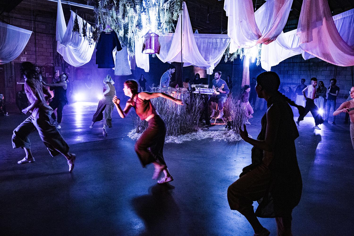 dancers running around musicians in a dark space with white drape hanging from the ceiling