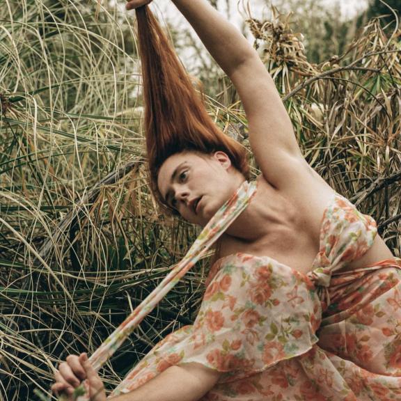 tess wearing a floral dress against green foliage, she has red hair, fair skin and is pulling both her dress string forward and hair up above her